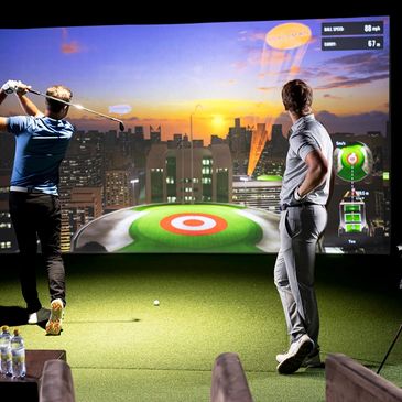 Two men playing golf on an indoor simulator with a cityscape sunset screen.