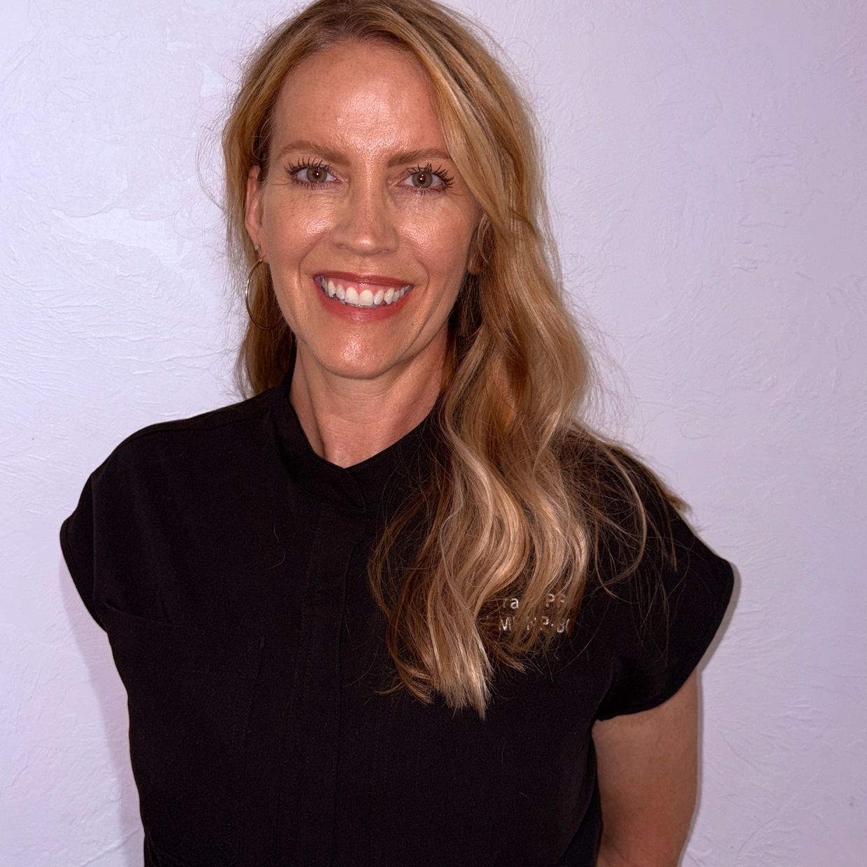 Smiling woman with long blonde hair wearing a black top against a light textured wall.