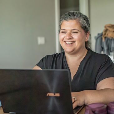 Woman smiling while working on a laptop at a wooden table.