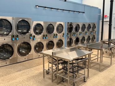 Modern laundromat with stainless steel dryers and folding tables.