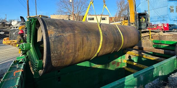 Large rusty cylindrical metal pipe being lifted by straps.