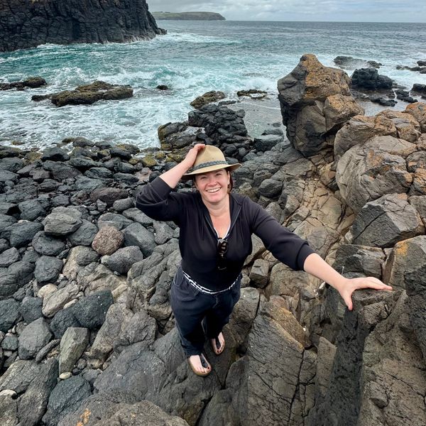 Smiling woman in hat climbing rocky coastline by ocean.