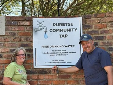 Two people smile beside a community tap offering free drinking water.