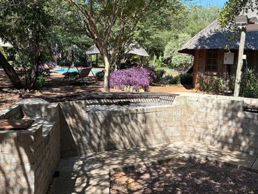 Outdoor stone barbecue area with trees, a pool, and a thatched-roof building in the background.