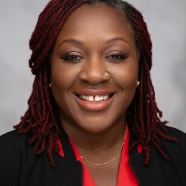 Smiling woman with red dreadlocks and a nose ring, wearing a red top and black jacket.