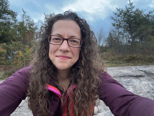 Woman with curly hair and glasses smiling outdoors on a rocky area.