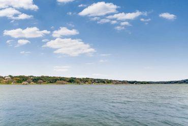 Calm lake under a partly cloudy blue sky with trees and houses on the far shore.