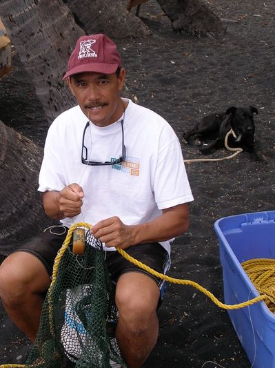 Man sitting on black sand beach fixing a fishing net with a dog in the background.