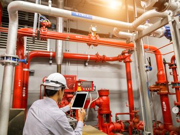 A technician in a helmet uses a tablet to monitor red industrial piping and valves