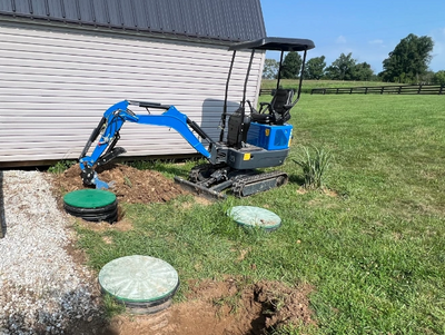 Mini excavator digging near a building with septic tank lids visible.