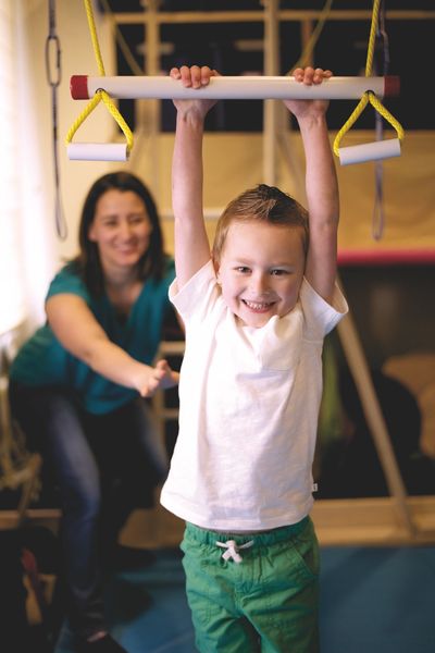 A child smiling while hanging on a swing bar with a therapist is pushing him from behind gently.