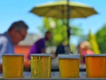 Four glasses of beer on a table with people and an umbrella in the background.