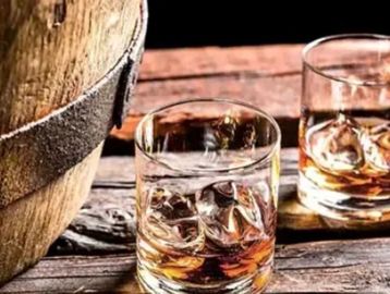 Two glasses of whiskey with ice on a wooden table beside a barrel.
