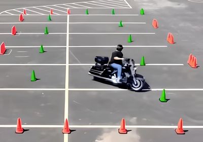 Person riding a motorcycle through a cone-marked course in an empty parking lot.