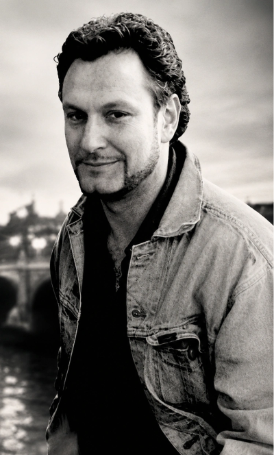 Man smiling in a denim jacket by a river.