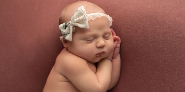 Sleeping baby with a white bow headband on a soft brown background.