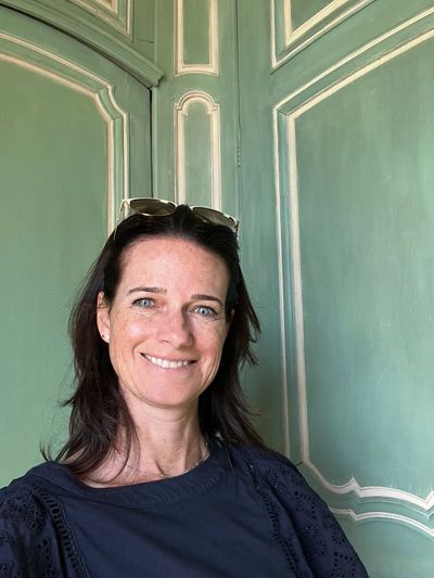 Smiling woman with sunglasses on her head, standing against a decorative green wall.