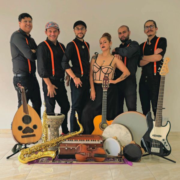 Banda musical con instrumentos variados y miembros vestidos de negro con tirantes naranjas.