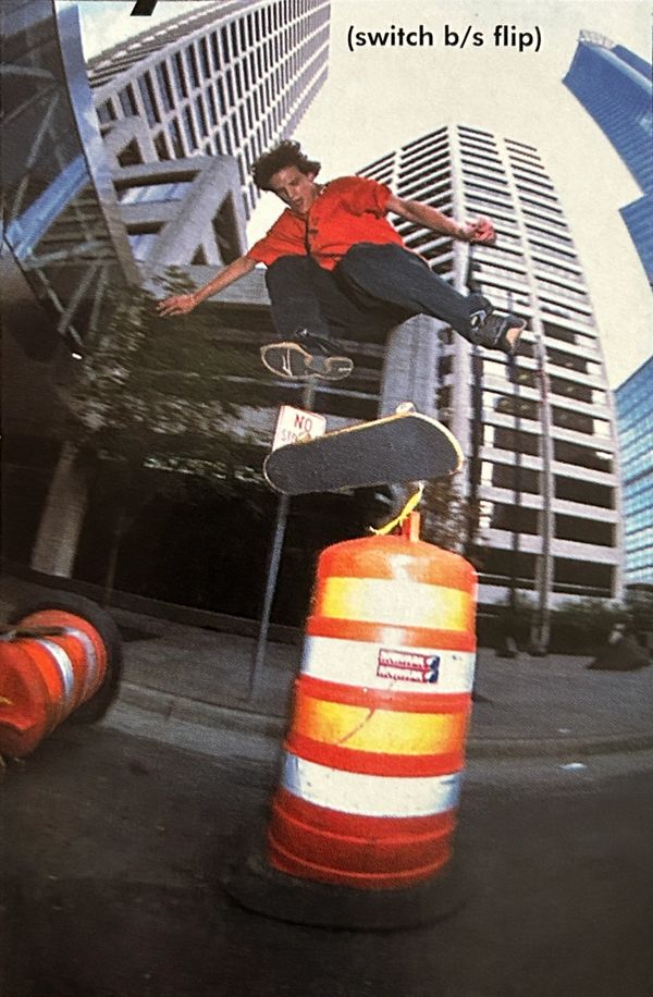 Skateboarder performs a switch backside flip over an orange traffic barrel in an urban setting.