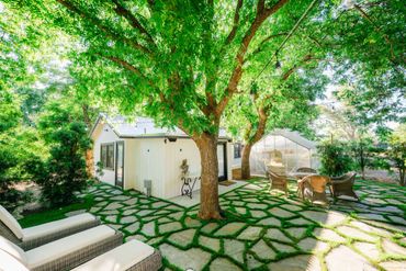 Sunny backyard with a white house, stone patio, large trees, and outdoor seating under string lights.