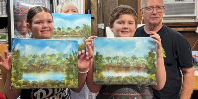 Two children proudly show landscape paintings with an older man behind them.