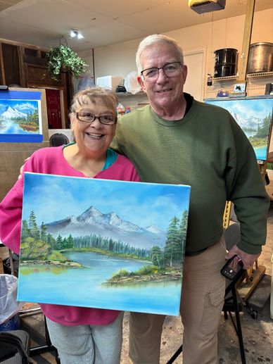 Two smiling seniors proudly display a landscape painting of mountains and a lake.