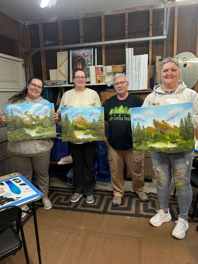 Four people proudly hold up paintings of mountain landscapes in a cozy indoor setting.