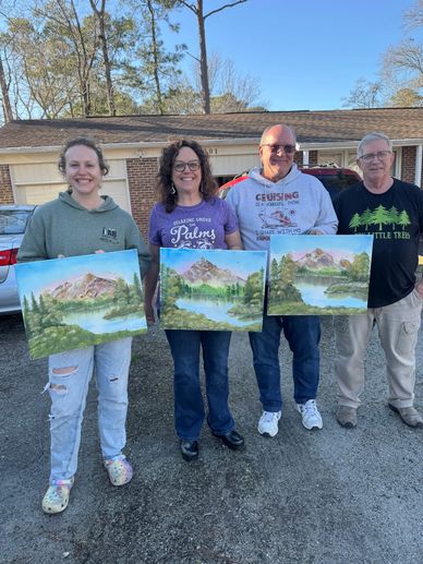 Four people proudly display their landscape paintings outdoors in front of a house.