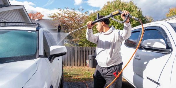 A man uses a pressure washer to clean a white vehicle in a driveway.