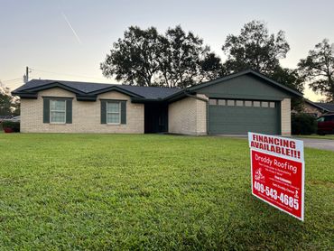 A beige brick house with green shutters and a roofing service sign on the lawn.