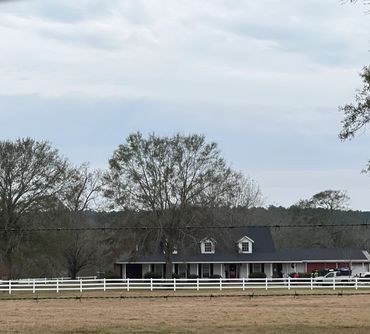 Rural house with white fence and large trees under a cloudy sky.
