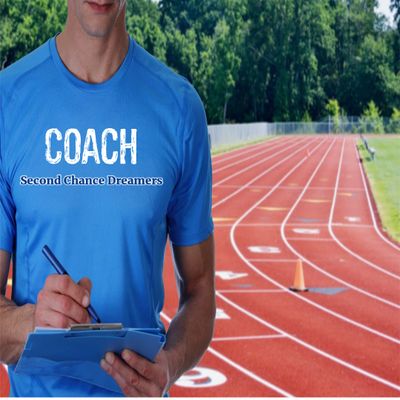 A coach in a blue shirt stands by a running track, writing on a clipboard.
