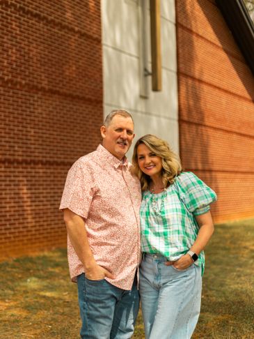 A smiling couple stands close together outside against a brick building.