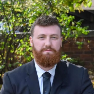 Man with red beard wearing a black suit and tie outdoors.