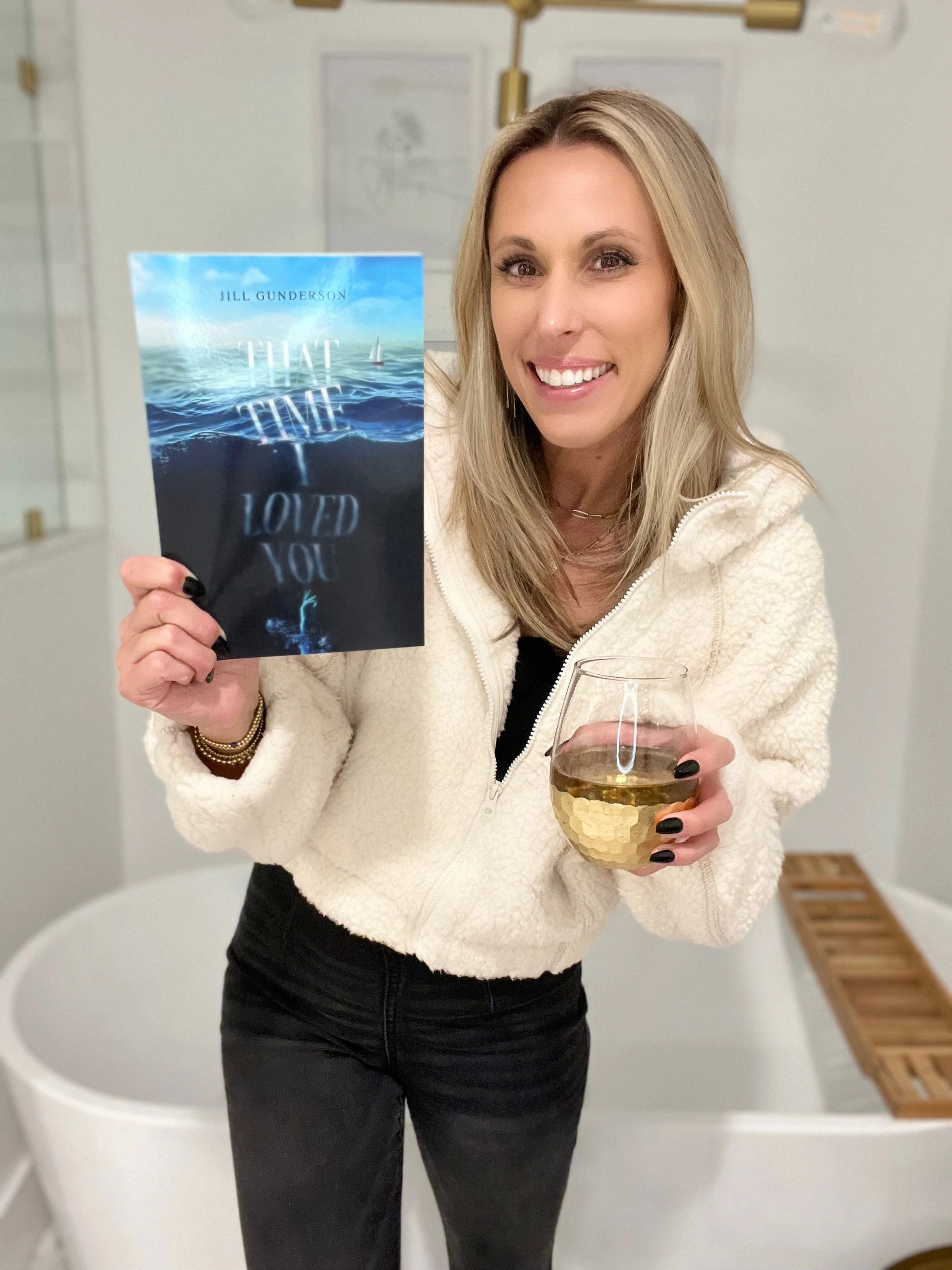 Jill is standing in her bathroom, holdering her book and a glass of wine.