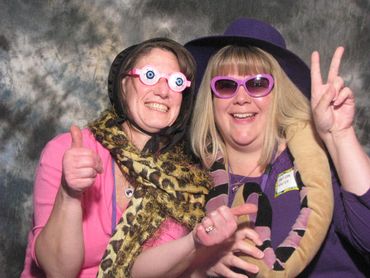 Two women wearing fun glasses and scarves, smiling and posing with thumbs up and peace sign.
