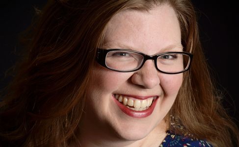A picture of Kelly, a woman with long, wavy red hair and dark framed glasses and a big smile.