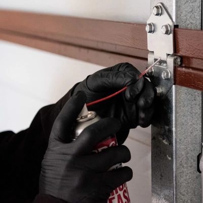 A gloved hand spraying lubricant on a garage door hinge