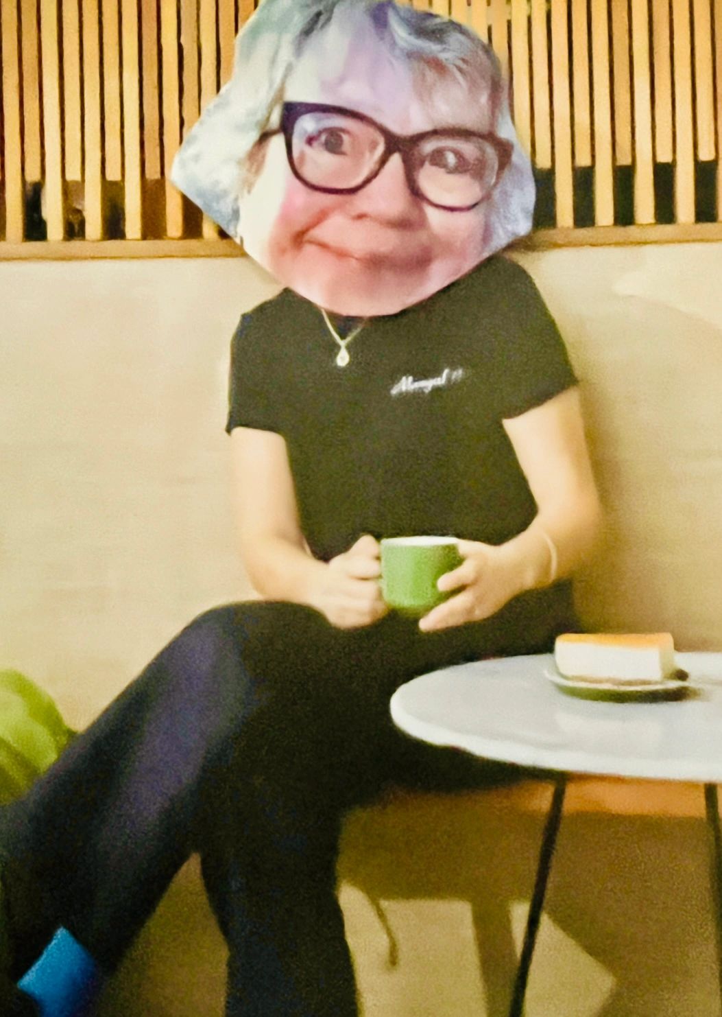 A Weir Sister with head clued on a model sitting at a table with a cup of coffee and cheese cake.