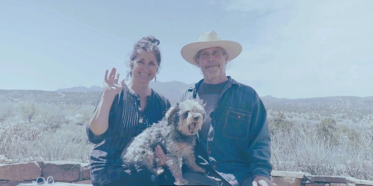 Smiling couple with a dog waving outdoors.