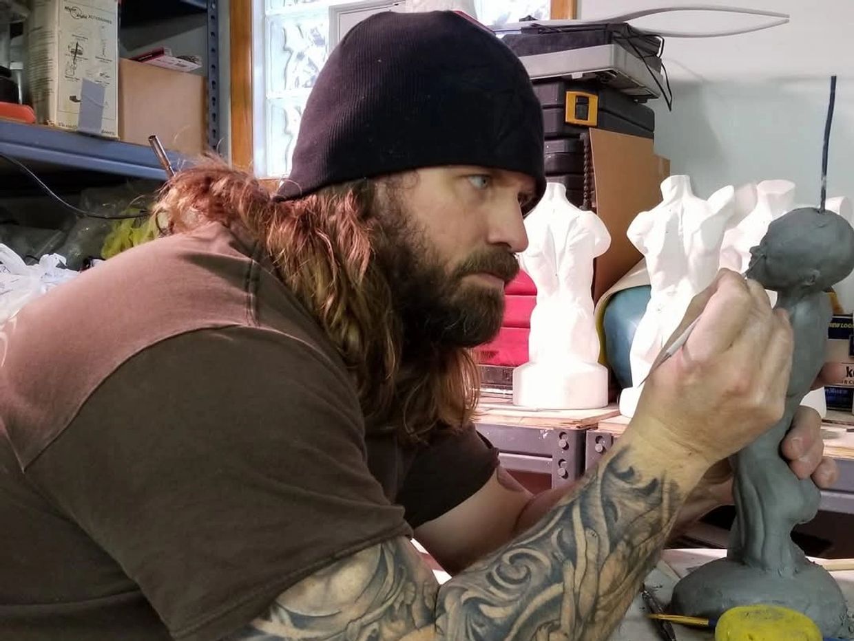 Tattooed artist sculpting a small figure with focus and precision.