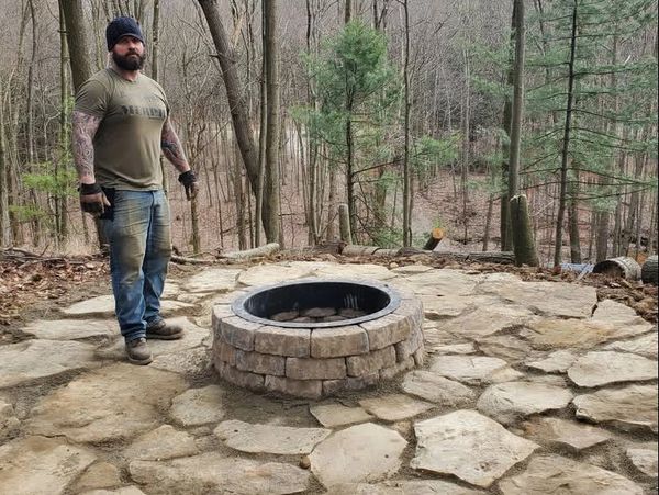 Man stands beside a newly built stone fire pit in a wooded area.