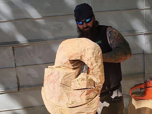 Man carves a large wooden sculpture outdoors using a chainsaw.