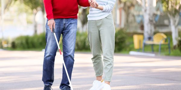 A visually impaired man walking with a white cane, guided by a woman holding his hand and a coffee cup.