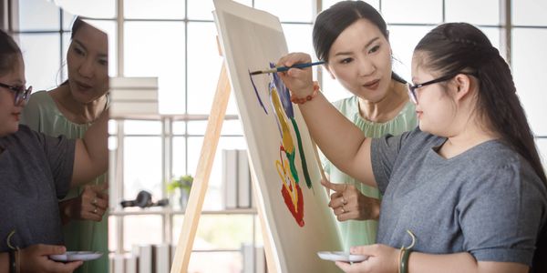 A teacher guides a young girl painting on a canvas in a bright room with large windows.