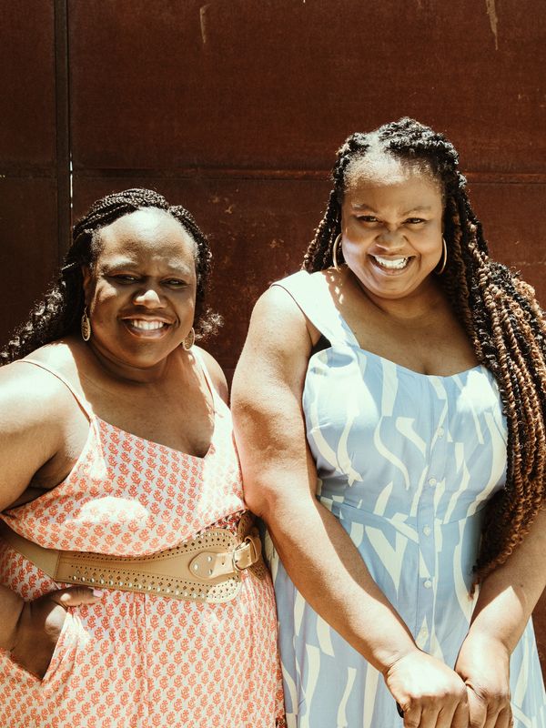 Two smiling women in summer dresses with braided hair standing outdoors.