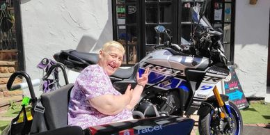 Smiling elderly woman in wheelchair near a parked motorcycle outdoors.