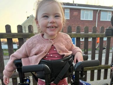Smiling toddler using a walker outside near a wooden fence.