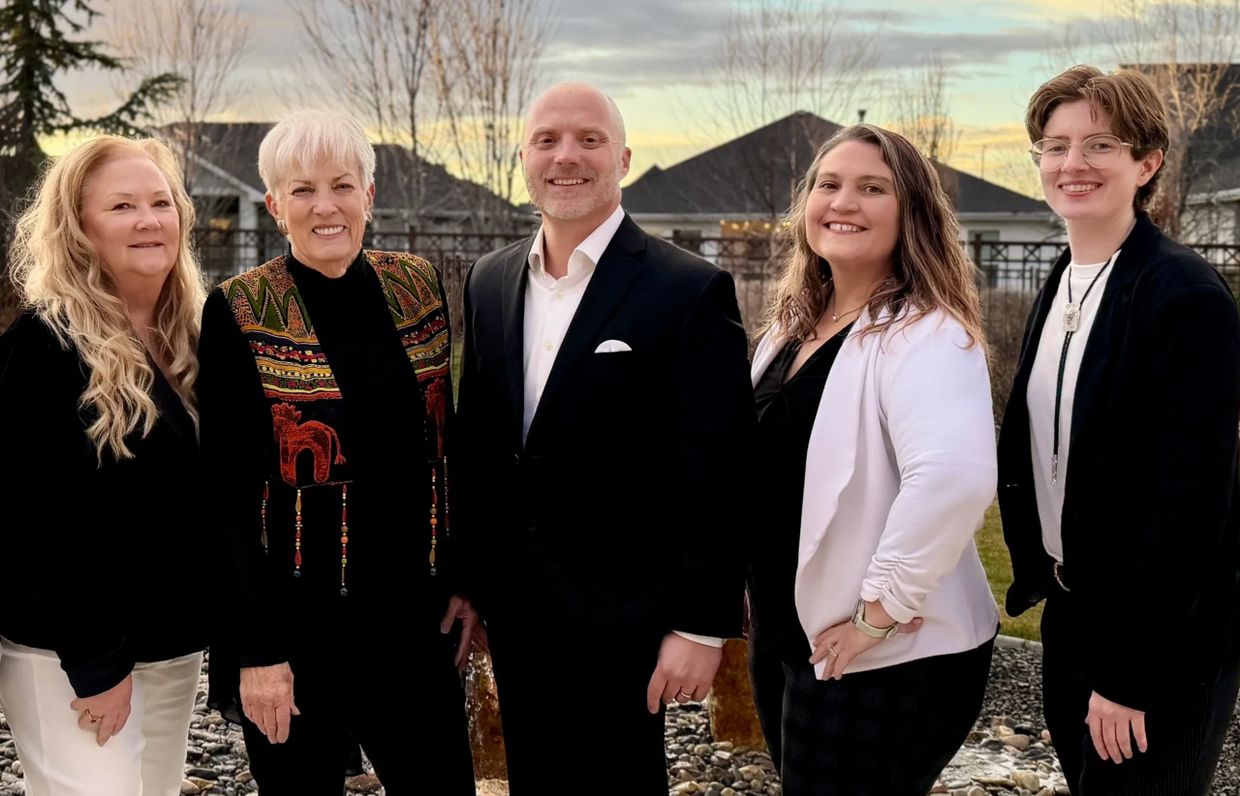 Five adults smiling outdoors in formal and casual attire during sunset.