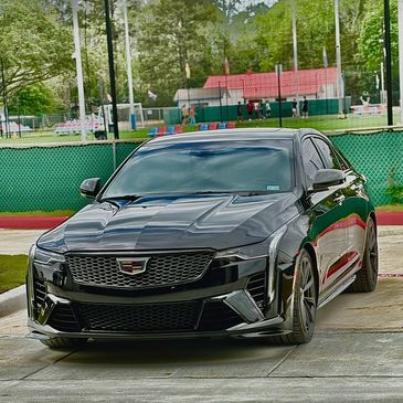 Sleek black Cadillac parked outdoors near a green fence and sports field.